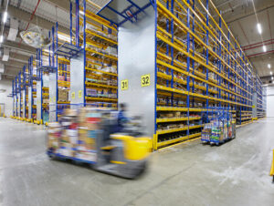 Mit hohem Tempo navigiert ein Kommissionierer durch die blau-gelben Regalzeilen und stellt Waren zusammen