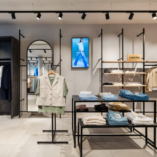 Flexible Wandregale mit Holzfachböden und schwarzem Metallrahmen ermöglichen eine markenbezogene Zonierung; digitale Screens ergänzen die Warenpräsentation