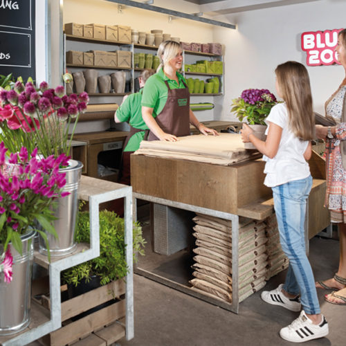 Der offen, frisch und stylish gehaltene Innenraum erinnert an einen stimmungsvollen Marktplatz. Für Theke, Warentische und Dekoration kam viel Holz zum Einsatz, und zwar massives, witterungsbeständiges Echtholz, das seine Wirkung nicht verfehlt. Hinter der Theke prangt eine große schwarze Fläche, beschriftet mit weißer Handschrift, im Tafel-und-Kreide-Look. Weitere Tafelflächen an den Wänden greifen das spontane Moment handgeschriebener Angebote auf – eine Assoziation an einen Marktstand. (Foto: Blume 2000)