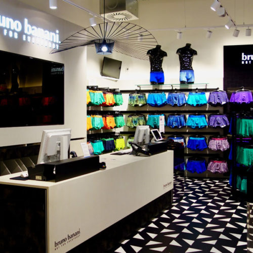 Unterstützt wird das Farbkonzept von einem Zusammenspiel verschiedener Haptiken und Oberflächen: Beschichtetes Metall und graues MDF stoßen auf kühlen Beton,  strukturierte Tapeten auf der einen Seite, seidenmatte Oberflächen auf der anderen bilden die dezente Ergänzung zur „lauten“ Bodengeometrie. Das Store-Konzept stammt von der Kraiss GmbH. (Foto: Bruno Banani)